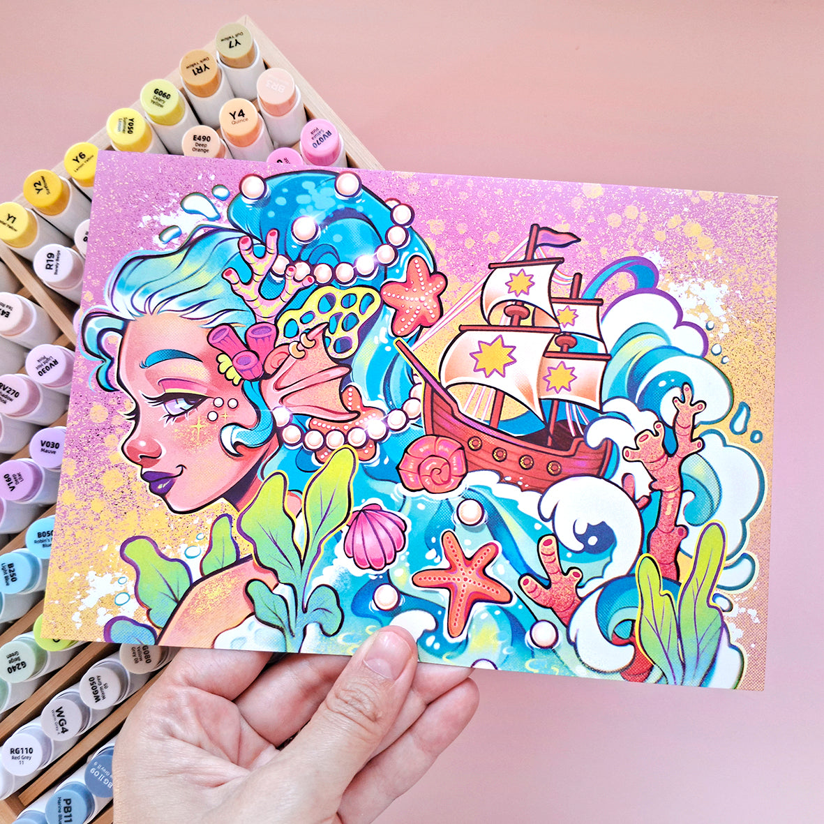 Colorful illustration of a mermaid and ship on a card held by a hand, with art supplies in the background.