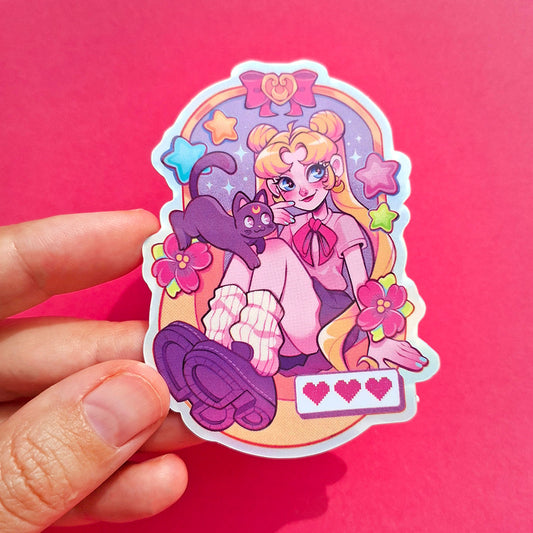 A hand holds a die-cut sticker featuring an anime-style blonde girl with twin buns, a black cat, flowers, and a purple shoe, set against a solid pink background.